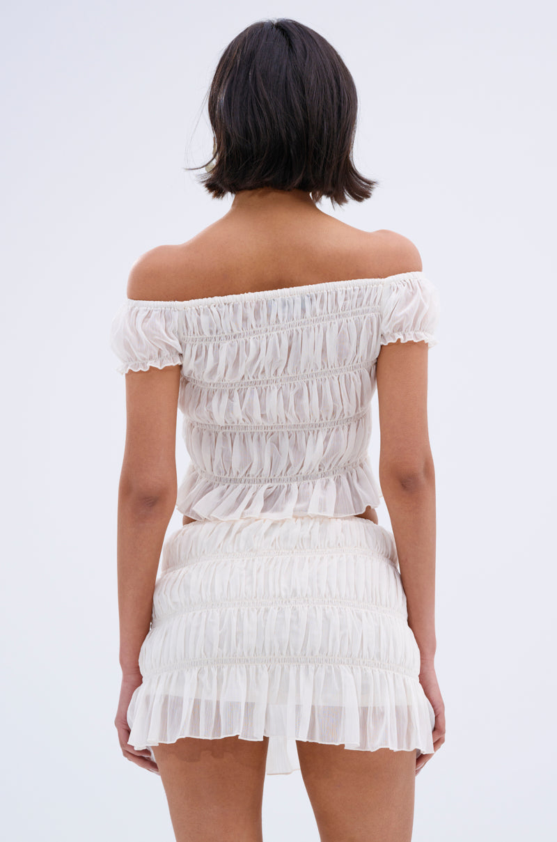 A woman with short dark hair faces away, wearing the FLIRT WITH ME OFF THE SHOULDER BLOUSE and a matching ruffled mini skirt crafted from sheer crepe fabric. The background is plain and light-colored.