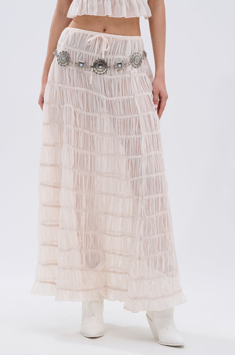 A person wears the JUST KEEP GOIN MAXI SKIRT—a sheer, pleated light pink maxi with seersucker detailing—styled with a decorative silver concho belt and white ankle boots against a plain white background.
