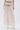A person wears the JUST KEEP GOIN MAXI SKIRT—a sheer, pleated light pink maxi with seersucker detailing—styled with a decorative silver concho belt and white ankle boots against a plain white background.