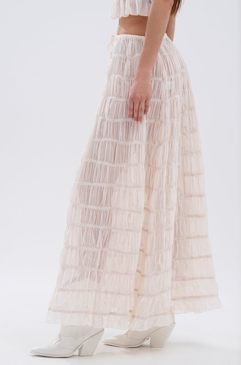 A woman in profile wears the JUST KEEP GOIN MAXI SKIRT—a long, sheer, textured beige skirt with seersucker and ruffles—paired with a matching cropped top and white pointed-toe boots against a plain white background.