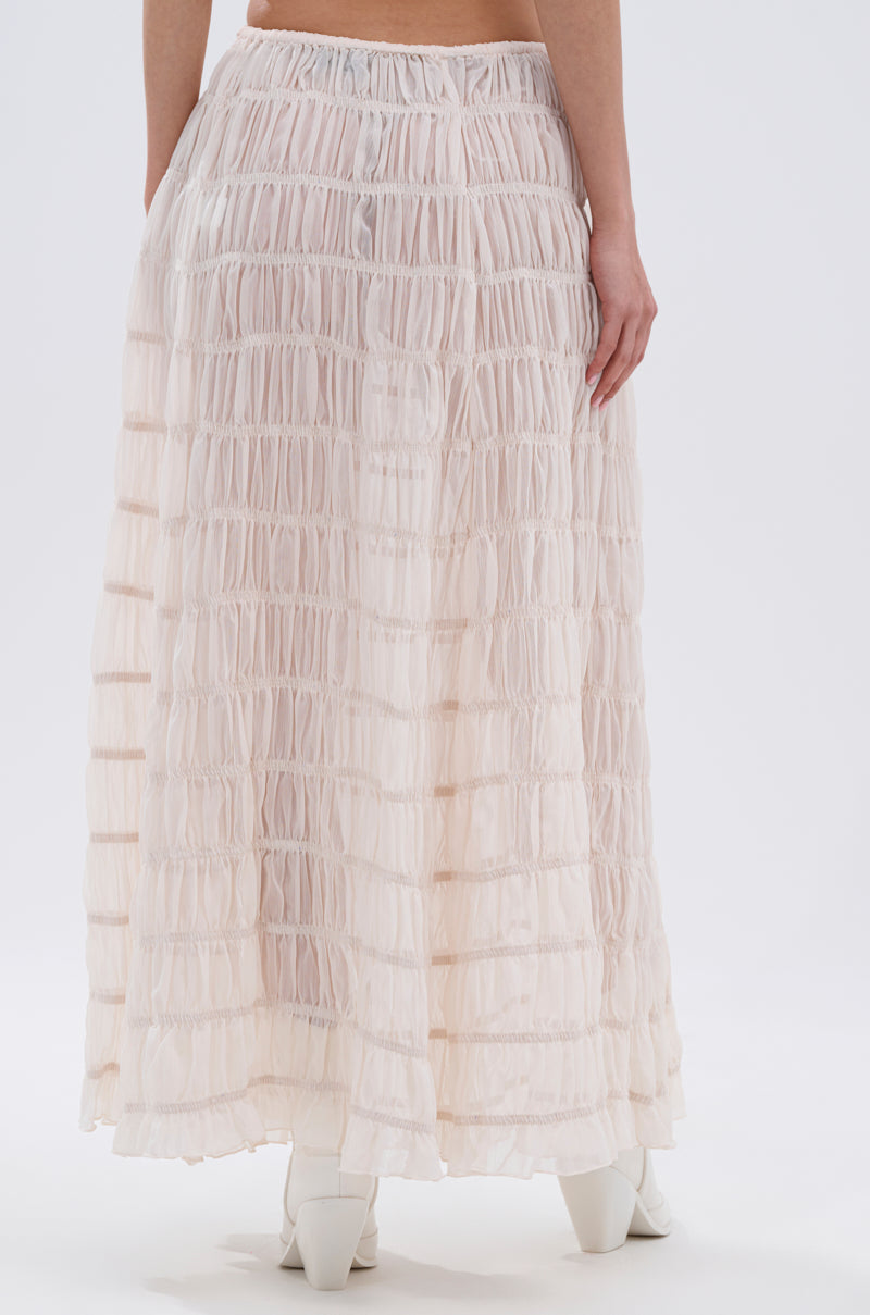A person stands with their back to the camera, wearing the JUST KEEP GOIN MAXI SKIRT in pale pink with seersucker detailing and a ruched texture. White heeled boots peek out from beneath the flowing, ankle-length skirt.