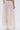 A person stands with their back to the camera, wearing the JUST KEEP GOIN MAXI SKIRT in pale pink with seersucker detailing and a ruched texture. White heeled boots peek out from beneath the flowing, ankle-length skirt.