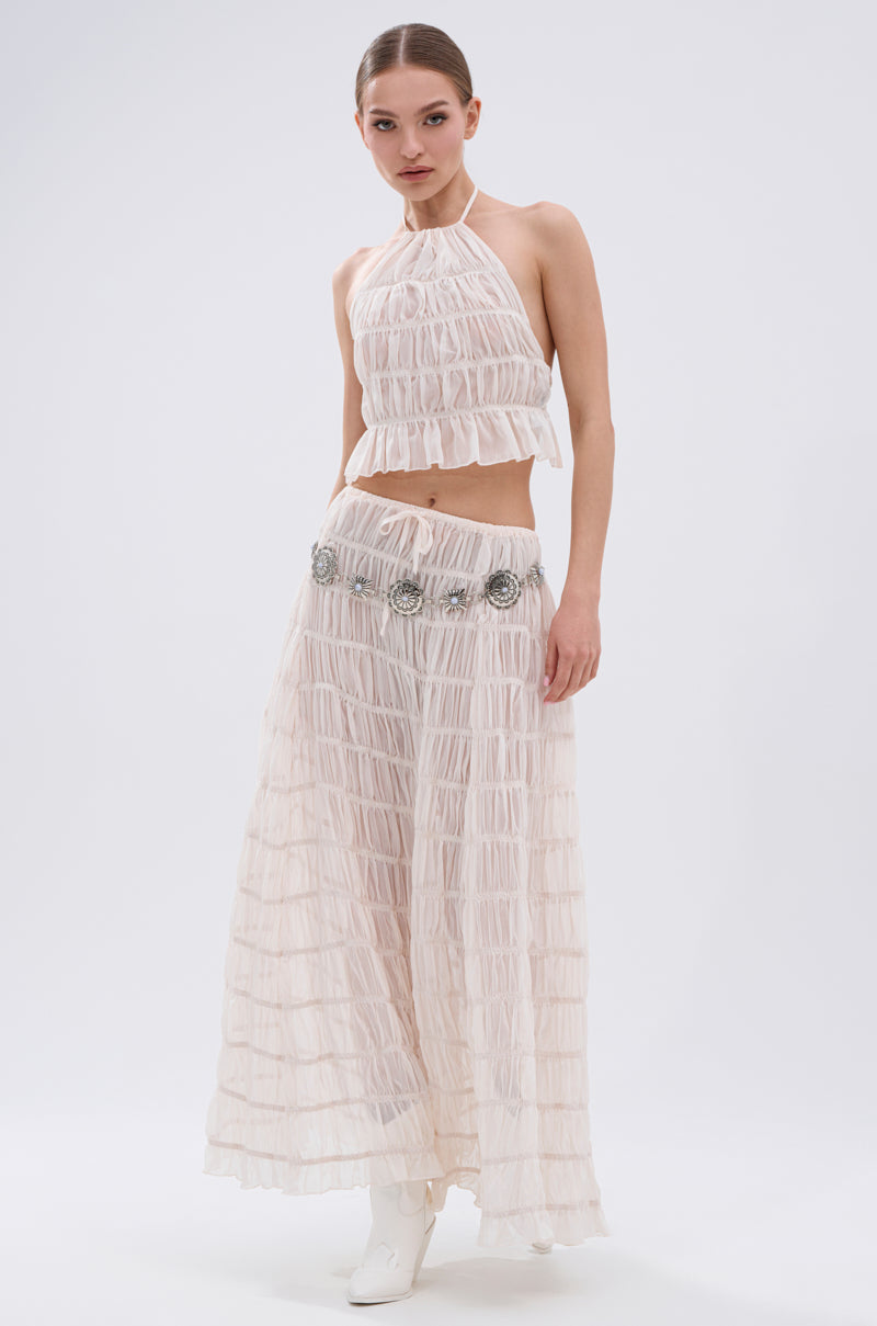 A woman models the JUST KEEP GOIN MAXI SKIRT—a light pink, sleeveless, halter-neck two-piece with seersucker detailing and pleated crop top—accessorized with a silver belt and white boots against a plain white background.