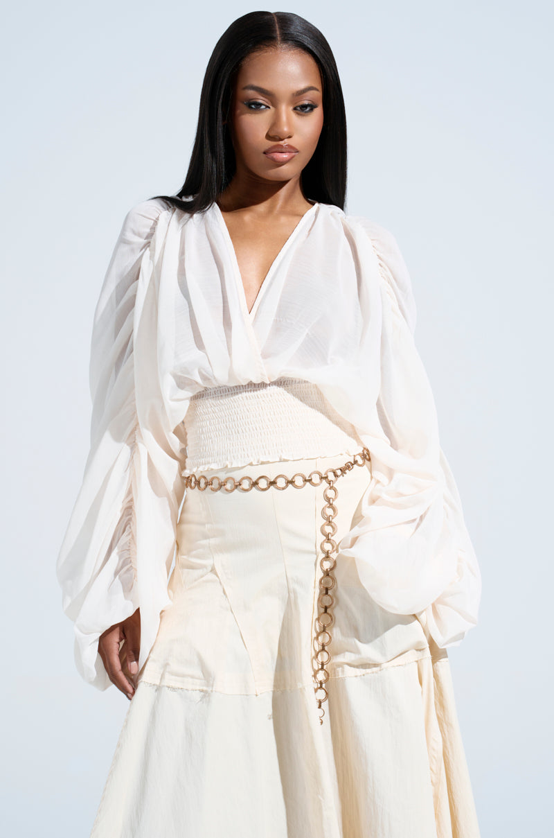 Someone with long, sleek hair wears a flowing white outfit featuring the JUST KEEP GOING BLOUSE with voluminous sleeves and a chain belt at the waist against a soft blue gradient background.