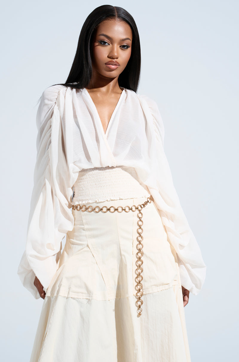 A woman with long hair dons a white, flowing outfit against a light backdrop. Her top, the "JUST KEEP GOING BLOUSE," features voluminous sleeves and a surplice neckline, elegantly paired with a high-waisted skirt and chain belt as she gazes confidently at the camera.