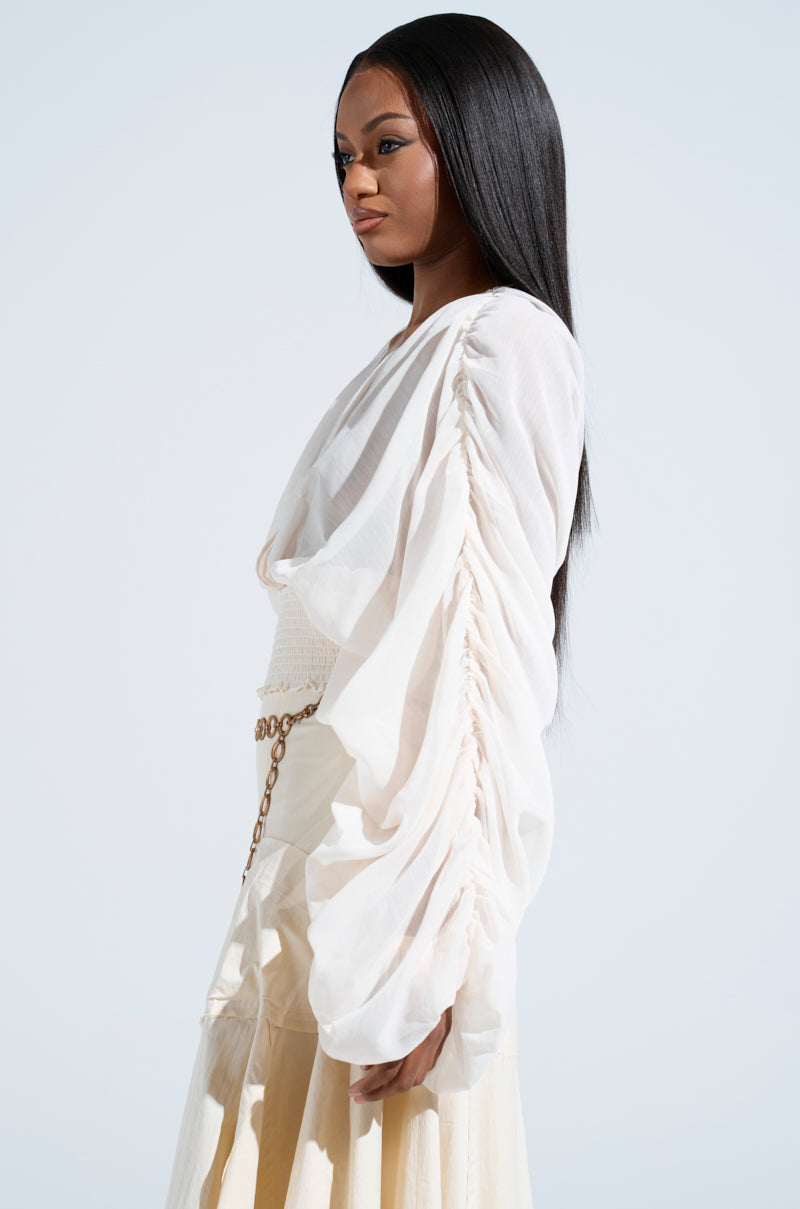 A woman with long straight hair wears the JUST KEEP GOING BLOUSE, a white chiffon top with ruched sleeves, paired with a beige skirt. She stands against a light blue background, gazing forward with a composed expression.