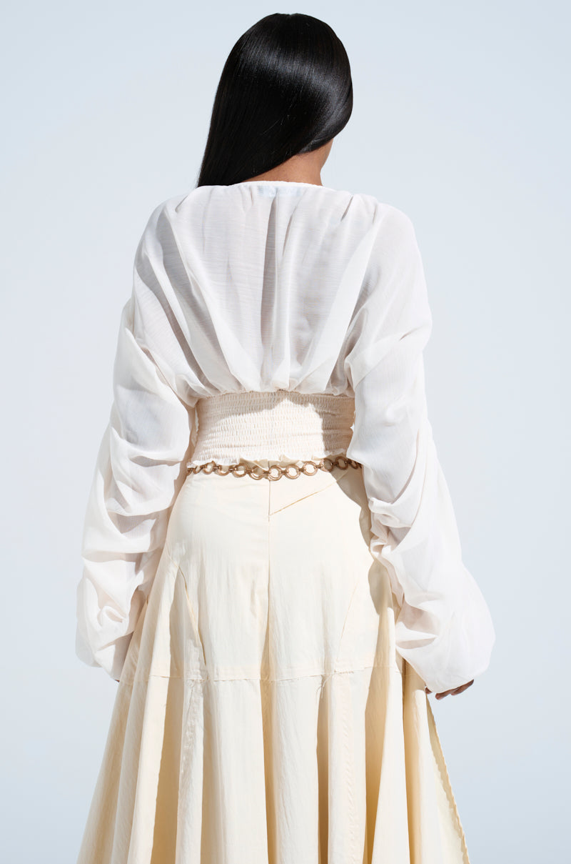 A person with long black hair is photographed from behind, wearing the JUST KEEP GOING BLOUSE and a cream skirt, highlighted by a beige woven belt. The light, neutral background enhances the elegant ensemble.