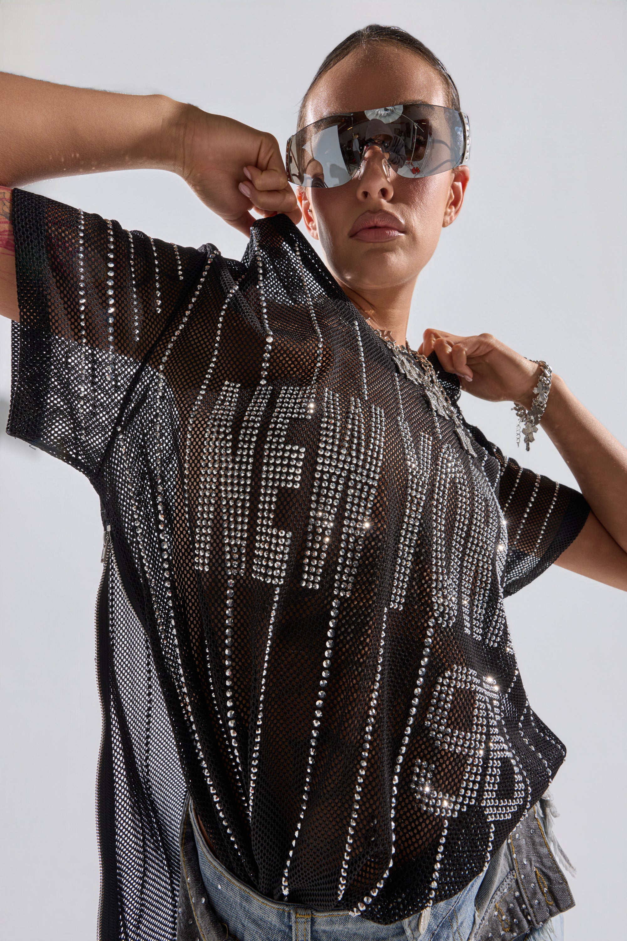A person confidently poses in the NEW YORK JERSEY—a bedazzled mesh top with rhinestones spelling "NEW YORK 95"—paired with large sunglasses, blue jeans, and bracelets, lifting one side of the shirt for added style. 0