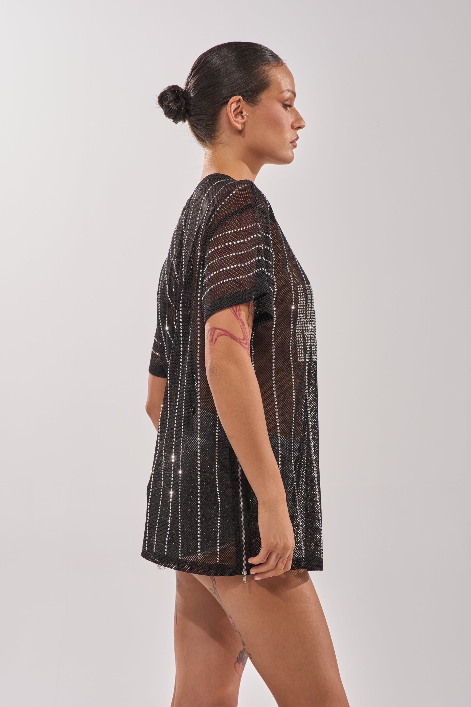 A woman with dark hair in a bun poses in profile wearing the NEW YORK JERSEY, a sheer black mesh top with rhinestone stripes over shorts. She has a visible arm tattoo and stands against a plain light background. 9