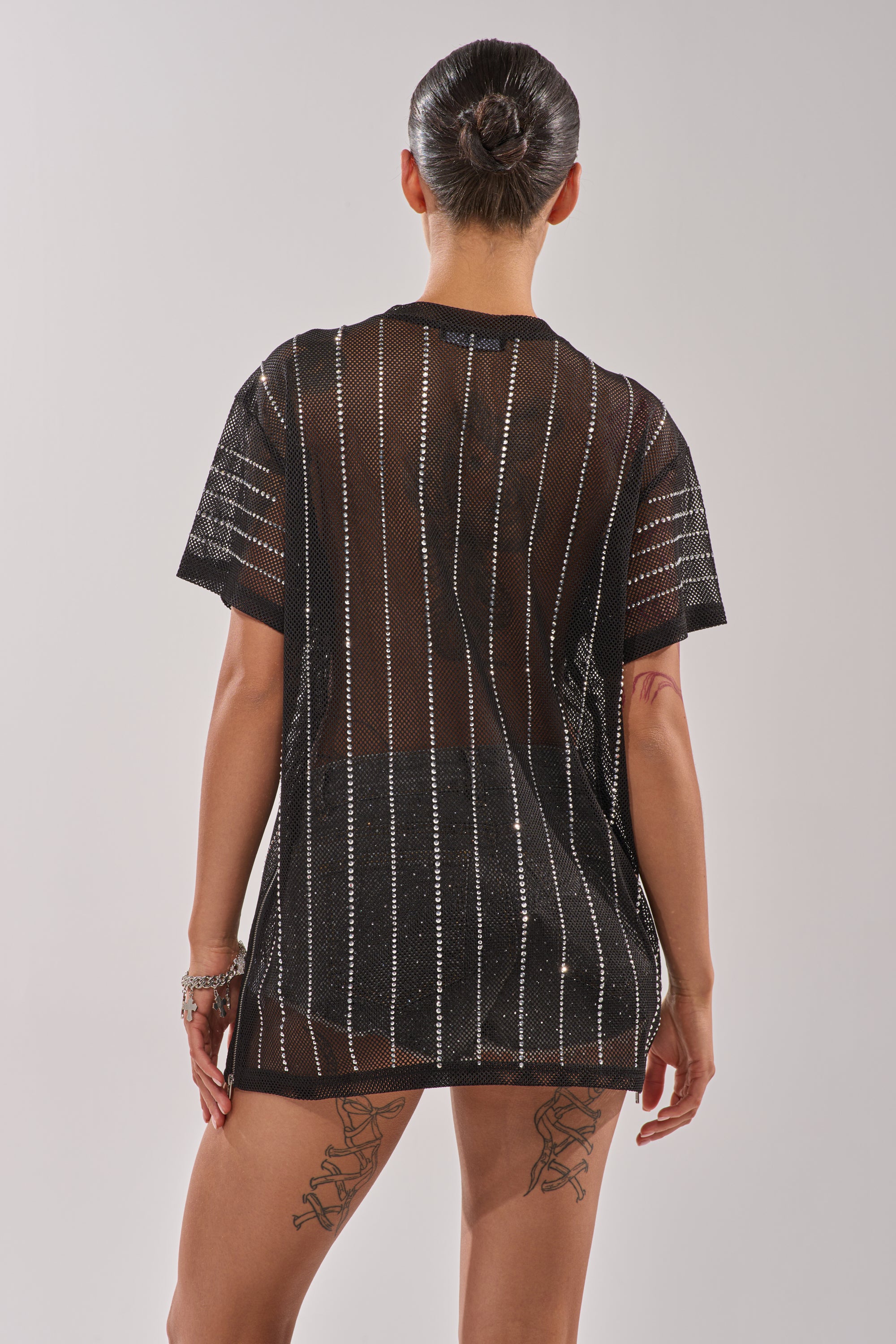 A person with their hair in a bun, facing away, wears the NEW YORK JERSEY—a sheer black short-sleeve mesh top with vertical silver stripes—showing tattoos on both legs and one arm against a plain light background. 10