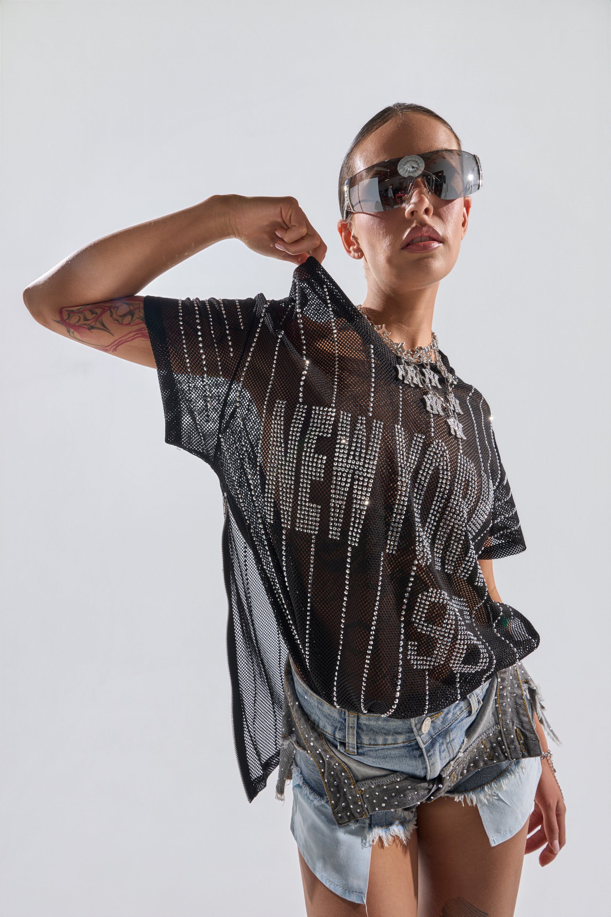 A person in sunglasses and a rhinestone-embellished NEW YORK JERSEY poses confidently, pulling on their shirt. They wear studded denim shorts, display arm tattoos, and have jewelry around their neck against a plain light background. 1