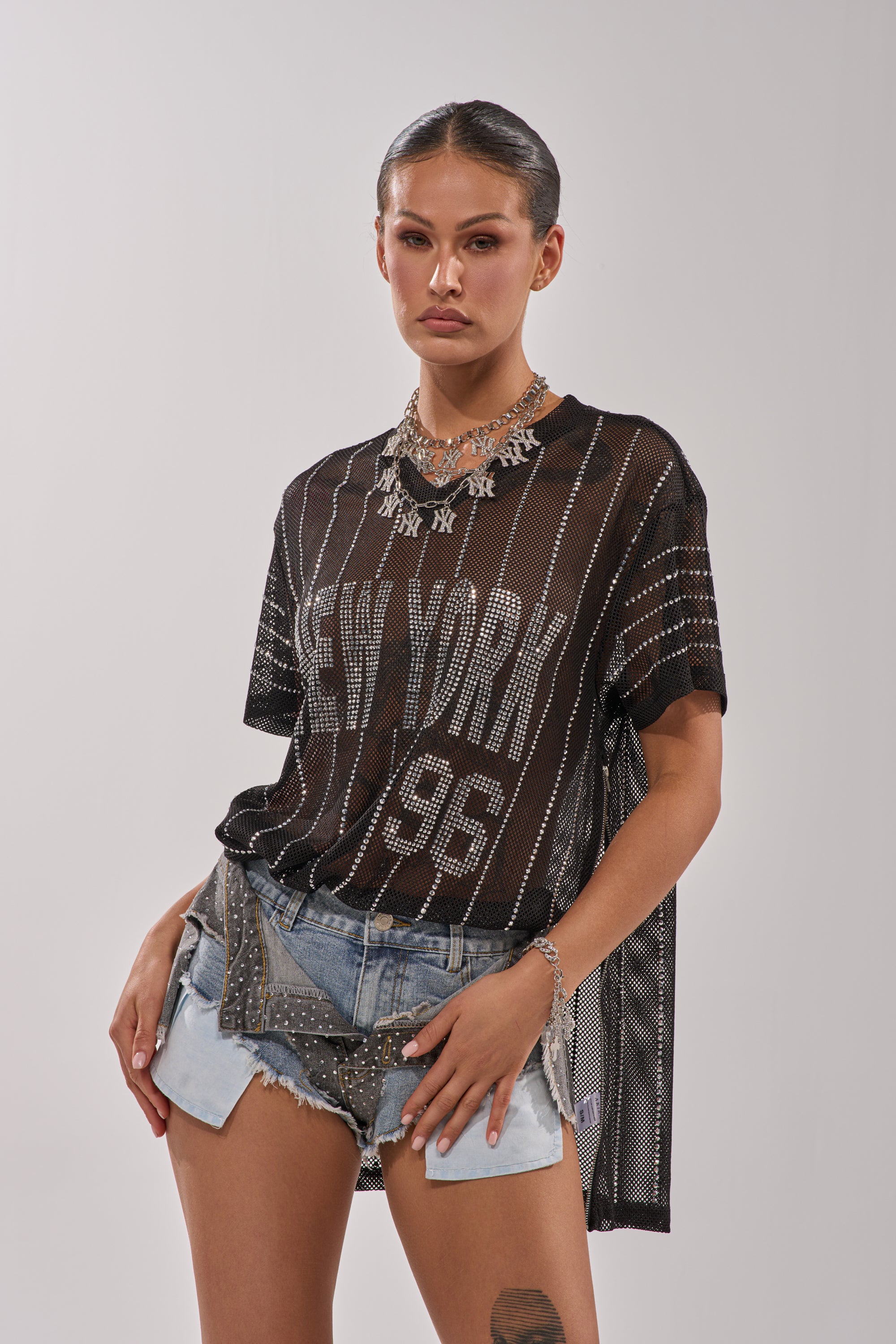 A woman confidently models the NEW YORK JERSEY, a sheer black oversized mesh top with "New York 96" detailing, styled over a black bra and paired with distressed studded denim shorts and layered necklaces. She maintains a neutral expression. 4