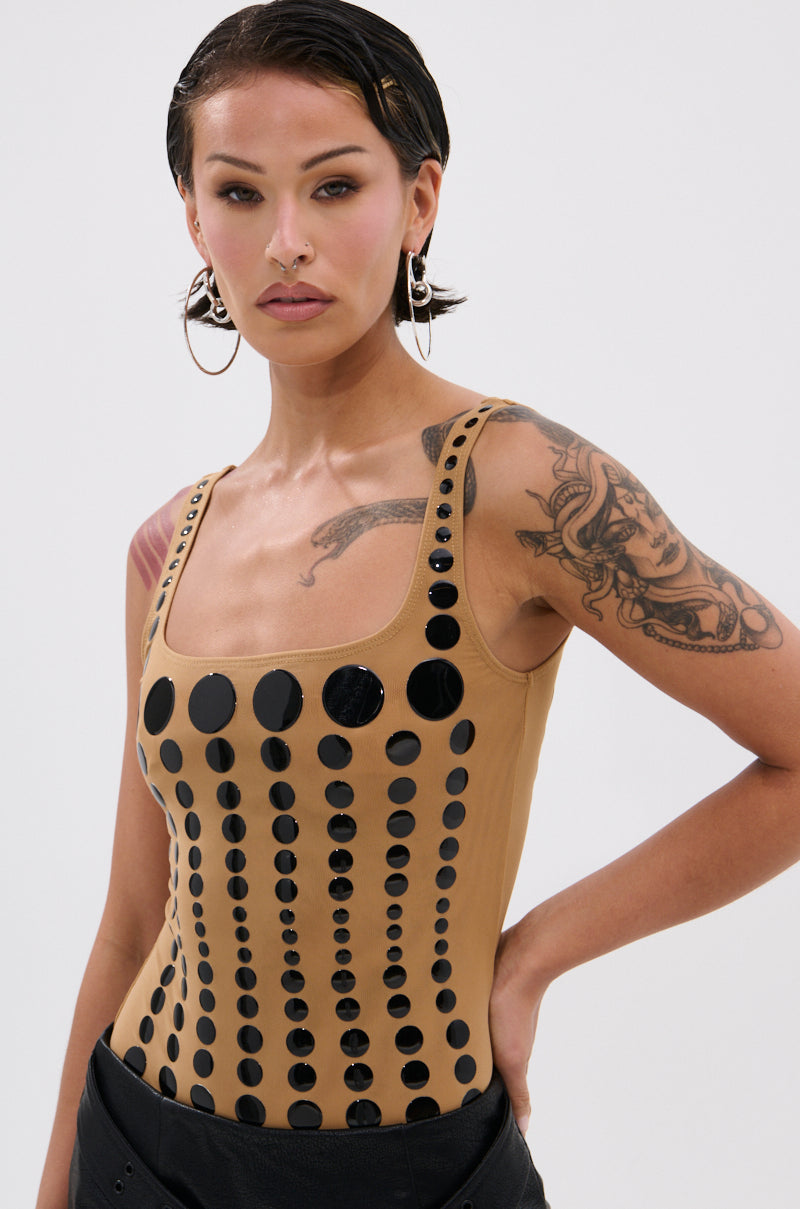A woman with short, slicked-back dark hair and tattoos models the NOT WHAT YOU THOUGHT POLKA DOT MESH BODYSUIT, featuring glossy black polka dots. She stands confidently with one hand on her hip against a plain white background.