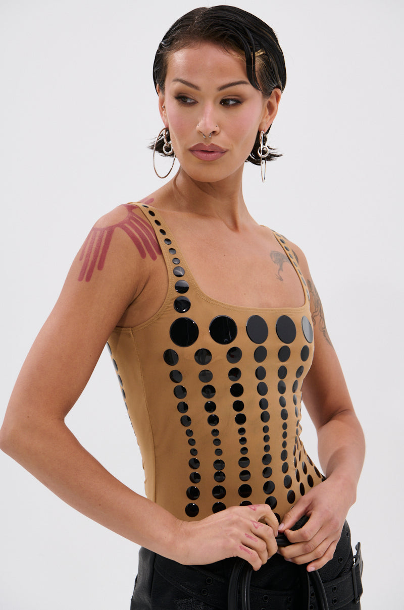 A person with short dark hair, hoop earrings, and arm tattoos models the NOT WHAT YOU THOUGHT POLKA DOT MESH BODYSUIT—a tan sleeveless top adorned with glossy black polka dots—against a plain white background.