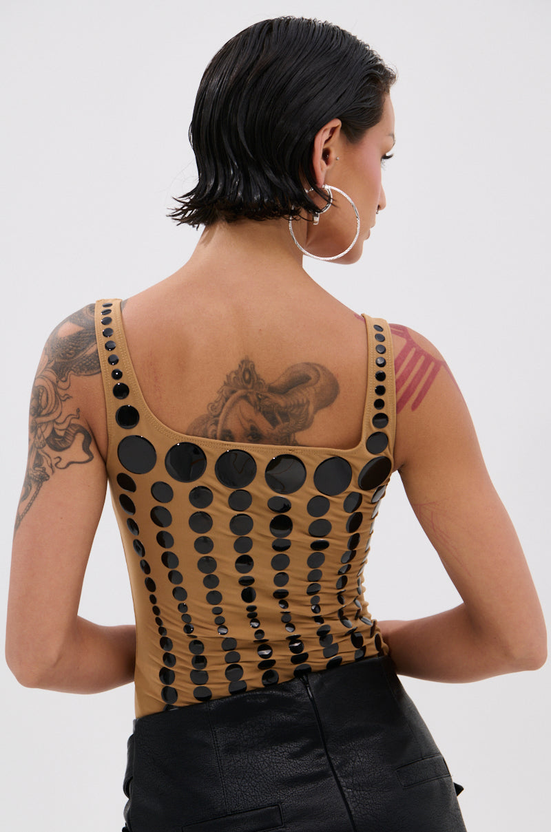 A person with short, wet-look hair wears the NOT WHAT YOU THOUGHT POLKA DOT MESH BODYSUIT with black high-waisted pants, hoop earrings, and showcases visible tattoos on their arms and shoulder.