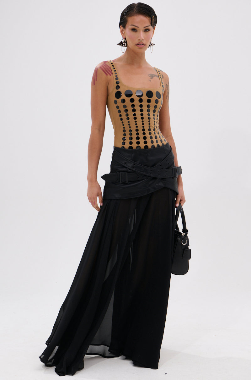 A woman models the "NOT WHAT YOU THOUGHT POLKA DOT MESH BODYSUIT," featuring glossy black polka dots, against a plain white background. She pairs it with a sheer skirt and holds a small black purse.