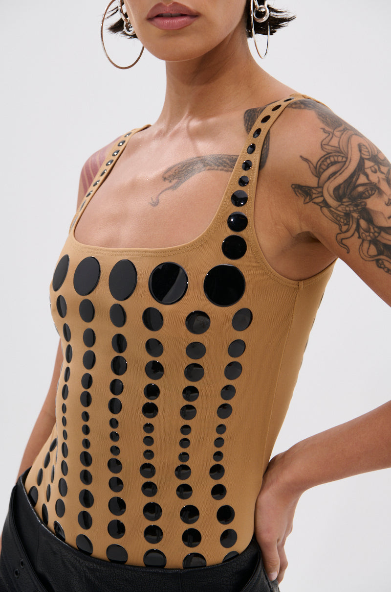A woman wears the NOT WHAT YOU THOUGHT POLKA DOT MESH BODYSUIT—a tan, sleeveless piece with glossy black polka dots—standing confidently with one hand on her hip, accessorized with hoop earrings and visible tattoos.