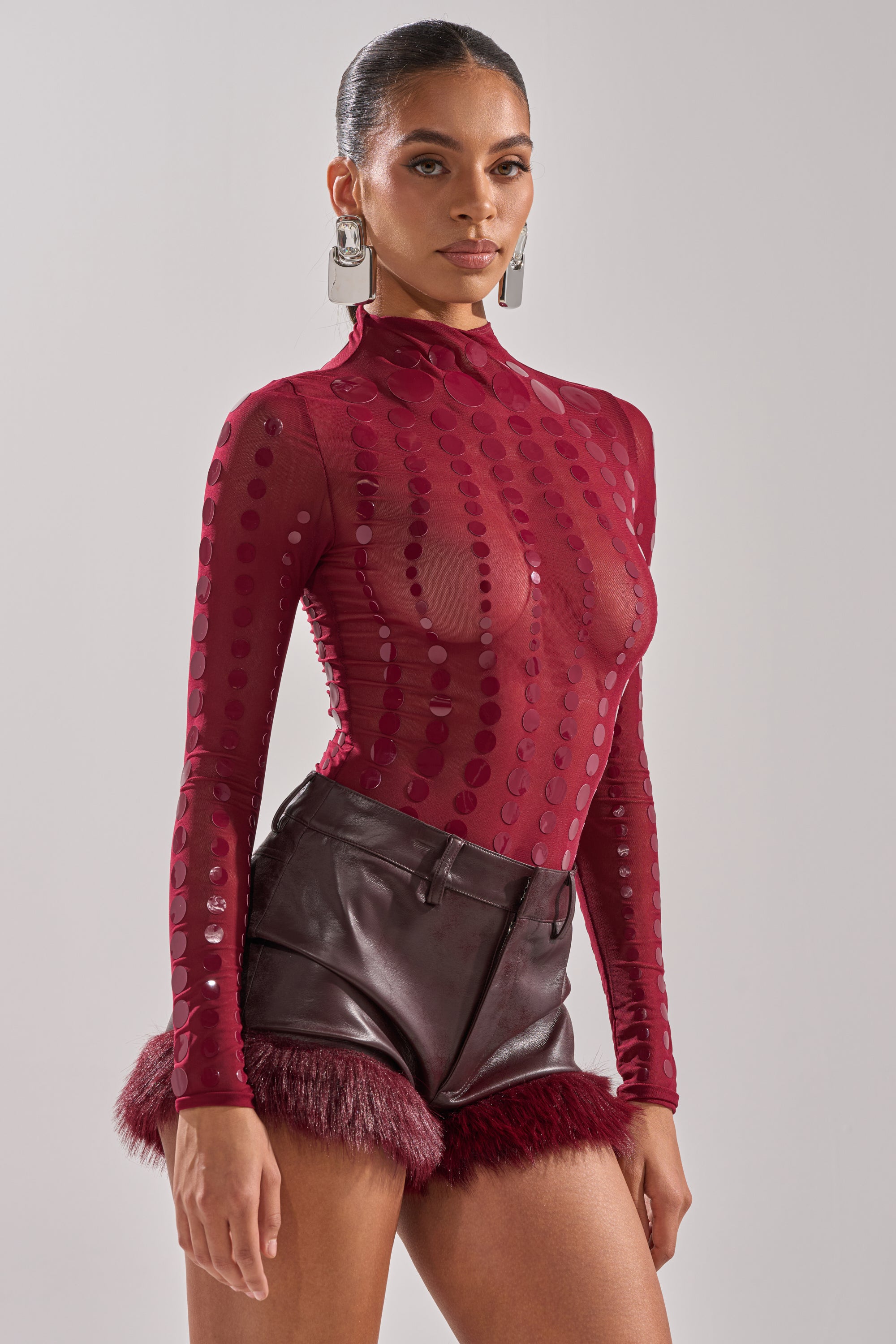 A woman stands against a plain background wearing the TRIPPY TYPE BODYSUIT, a sheer mesh style with long sleeves and polka dots, paired with high-waisted burgundy leather shorts with fur trim. She has statement earrings and her hair is pulled back.