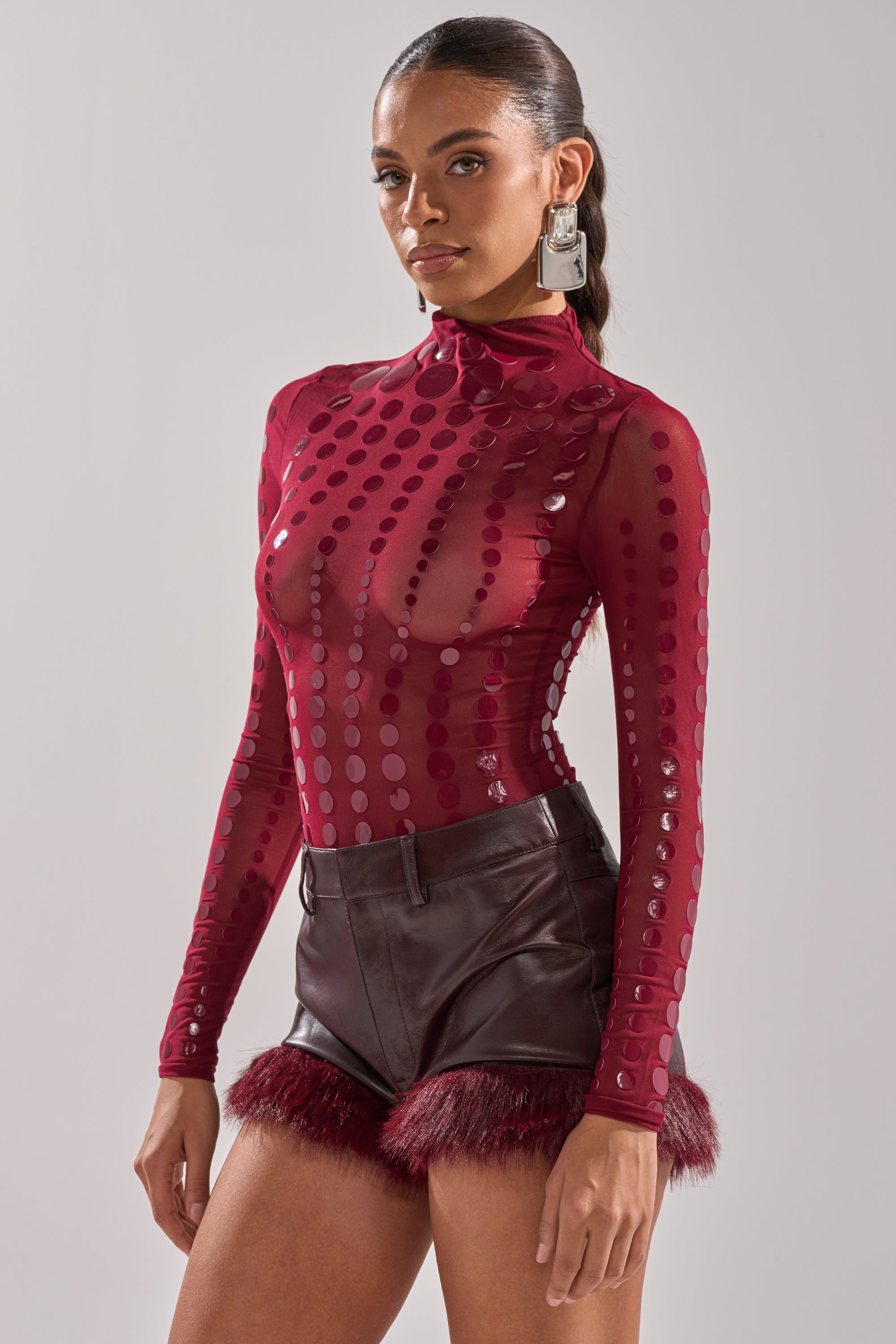 A woman wears the TRIPPY TYPE BODYSUIT, a sheer mesh maroon top with shiny polka dots, paired with high-waisted dark leather shorts trimmed in maroon faux fur. She adds large square earrings and keeps her hair pulled back.