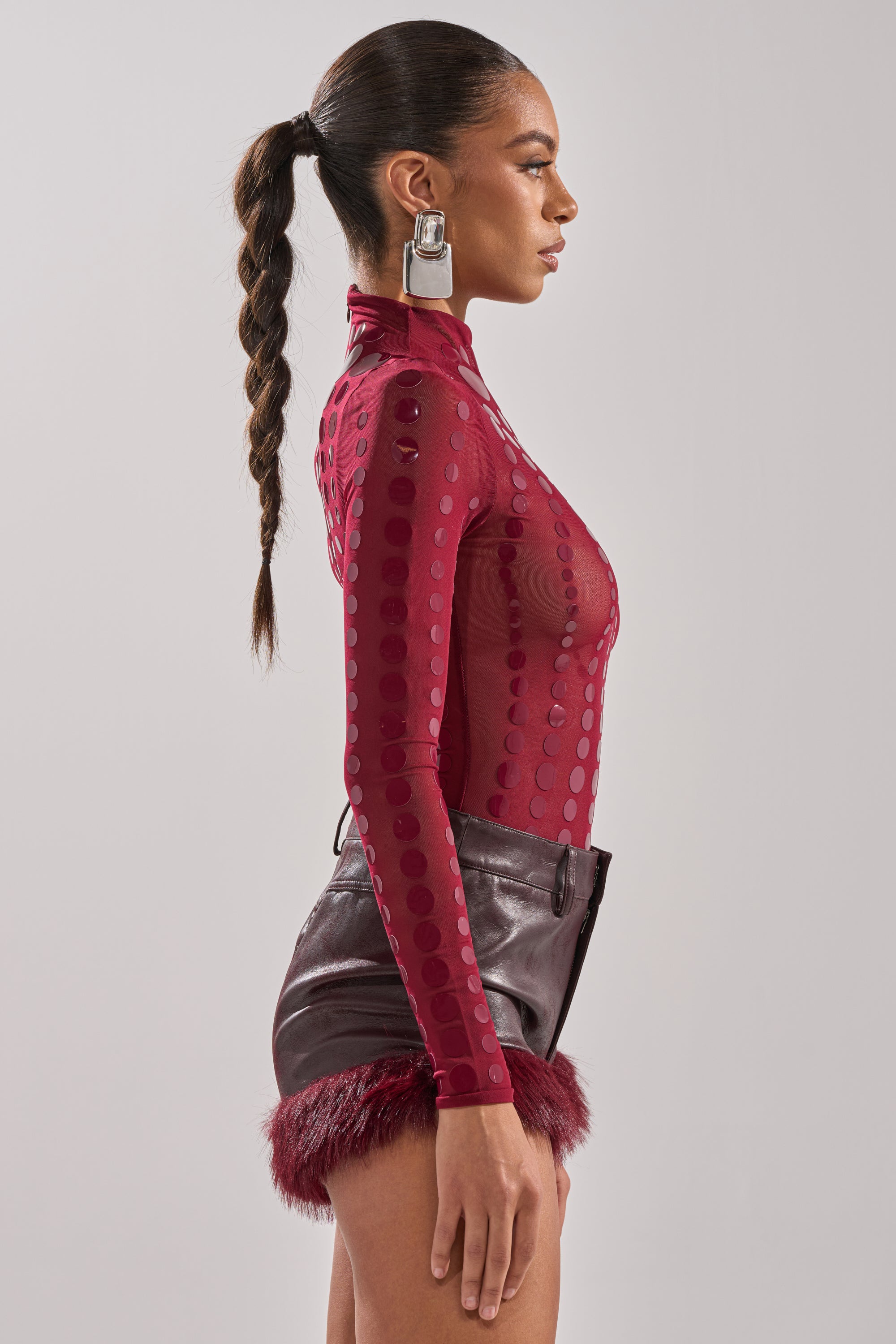 A woman stands in profile against a plain background, wearing the AKIRA Label TRIPPY TYPE BODYSUIT—a sheer mesh piece with red polka dots—paired with high-waisted dark red feather-trim shorts, large silver square earrings, and a long braided ponytail.