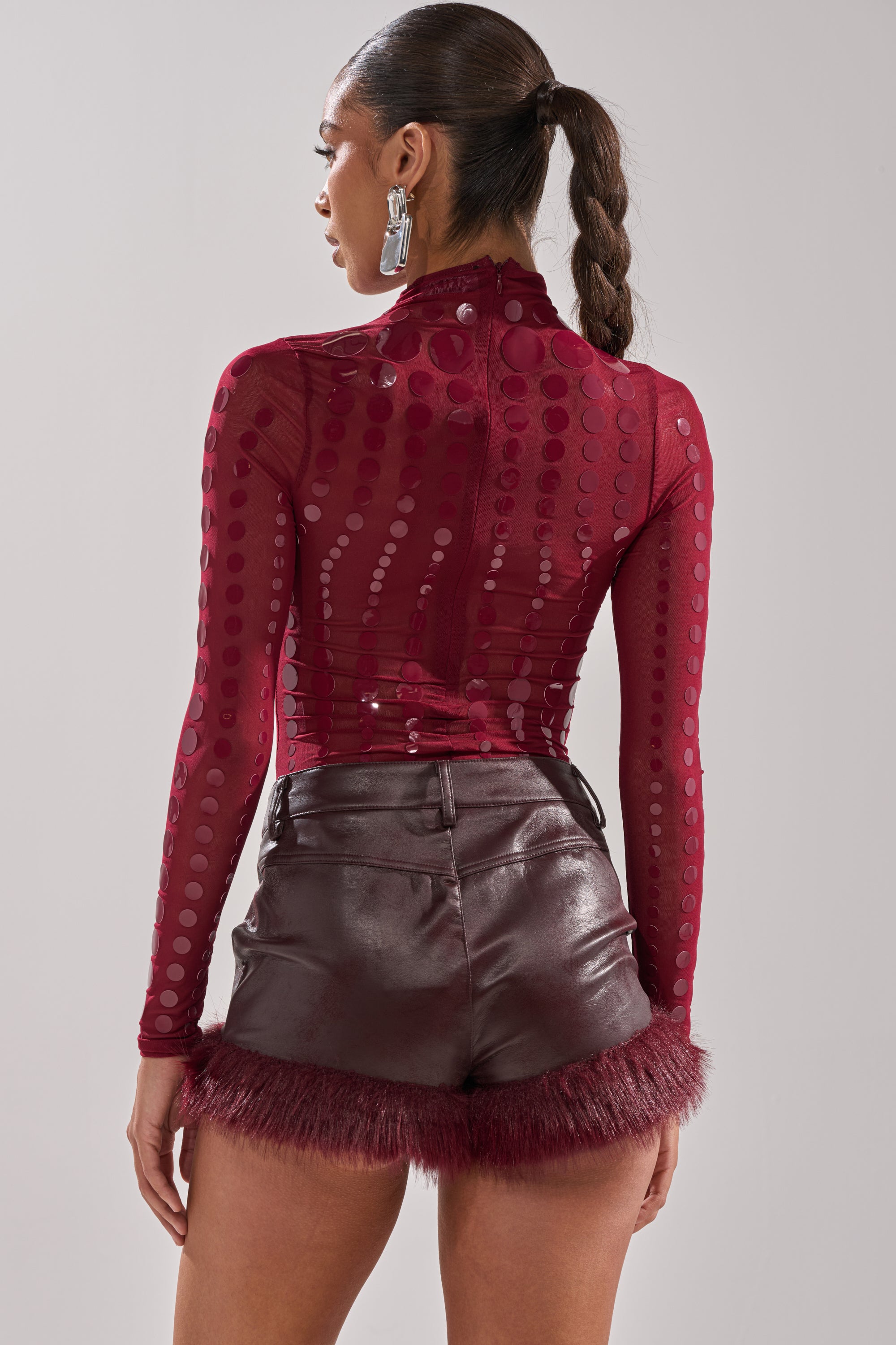 A woman with a long braided ponytail is seen from the back, wearing the AKIRA Label TRIPPY TYPE BODYSUIT in maroon polka-dots and high-waisted burgundy faux leather shorts with maroon feather trim, finished with statement earrings.