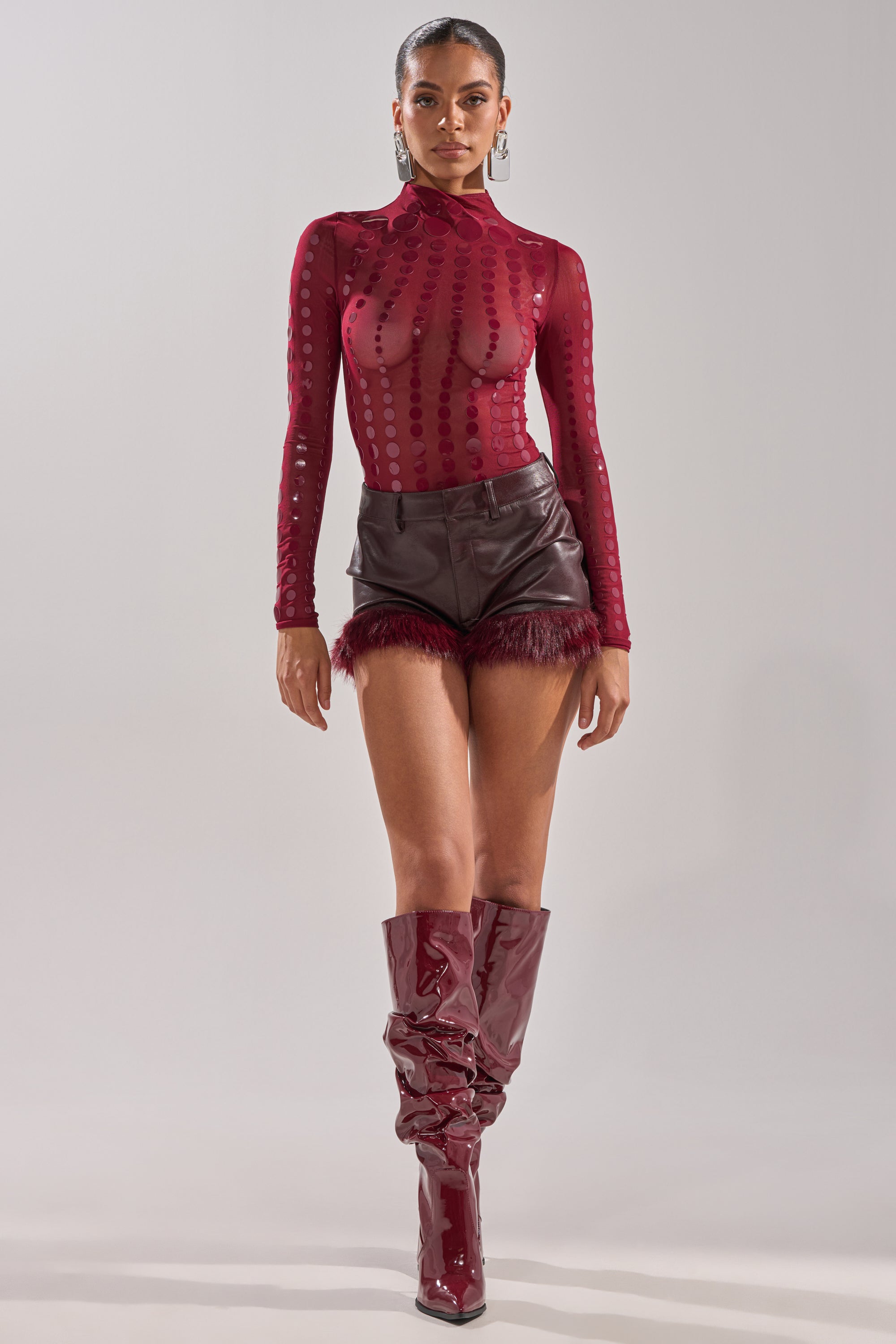 A woman walks confidently toward the camera wearing an AKIRA Label TRIPPY TYPE BODYSUIT, high-waisted maroon leather shorts with feather trim, and slouchy, shiny maroon knee-high boots.