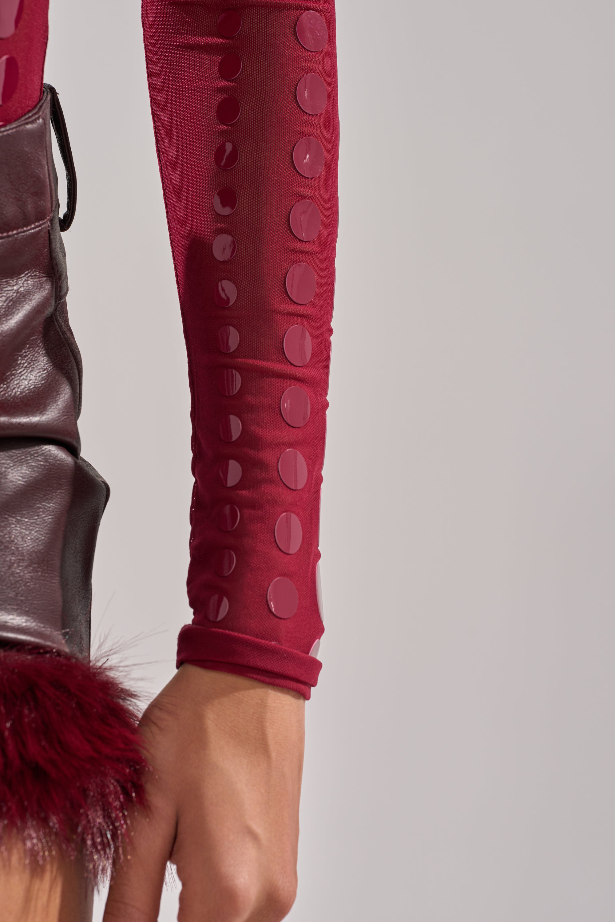 A person’s arm is shown wearing the TRIPPY TYPE BODYSUIT, featuring sheer mesh and raised dot patterns, paired with a leather garment with red faux fur trim, all set against a plain light background.