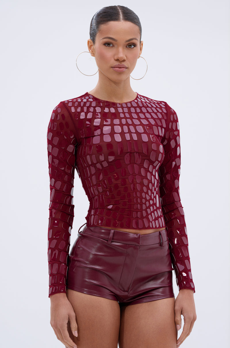 A woman with slicked-back hair and large hoop earrings models the CAN SEE RIGHT THROUGH YOU MESH LONG SLEEVE TOP in burgundy, paired with matching high-waisted leather shorts, standing against a plain light background. 2