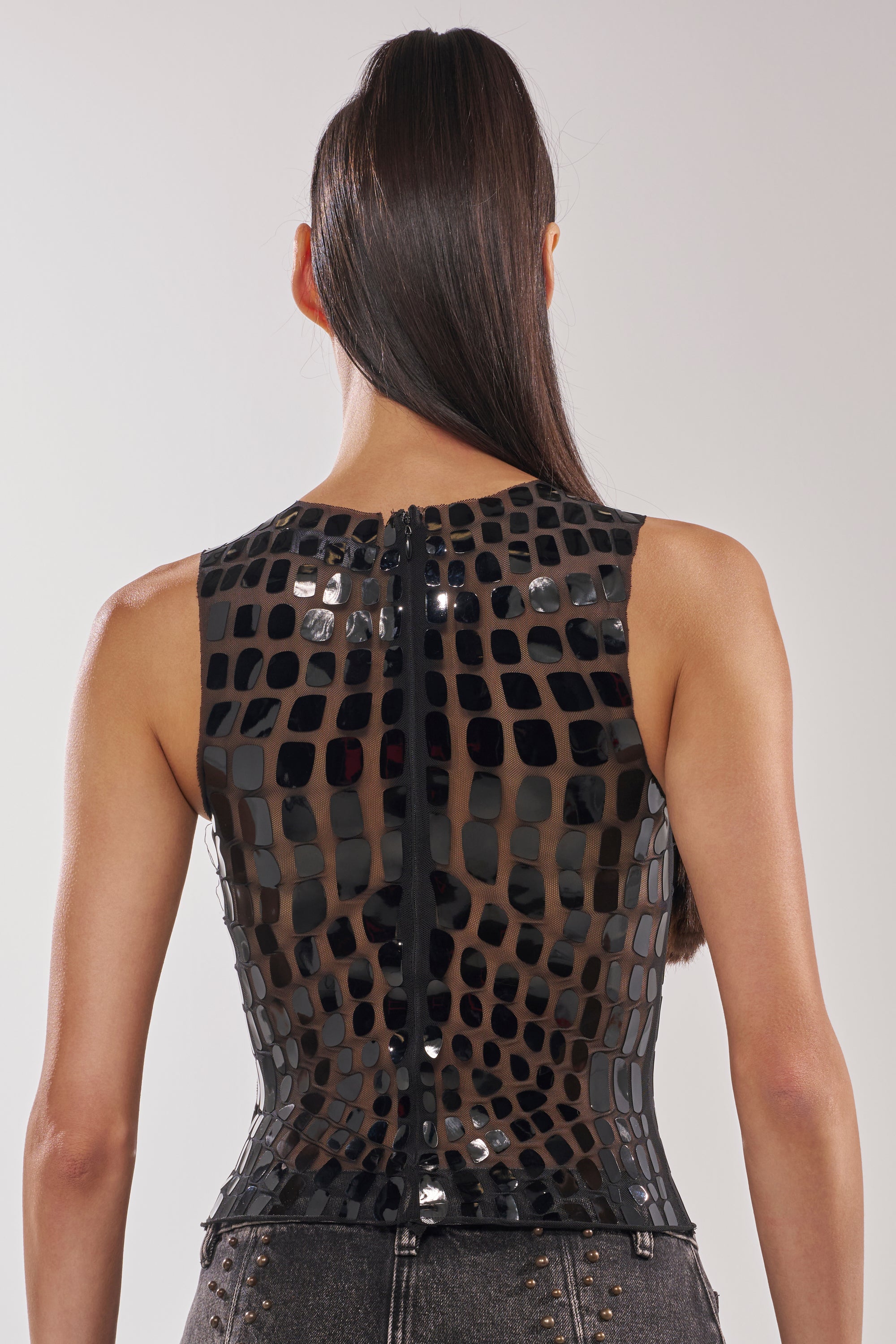 A woman with long, straight dark hair is seen from the back wearing the SEE THROUGH ME MESH TANK TOP—a black mesh tank with shiny patches—paired with high-waisted dark jeans adorned with metal studs. 4