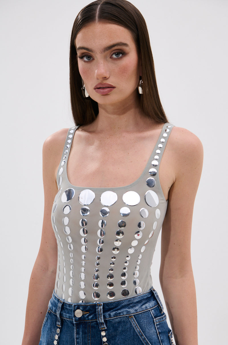 A woman with straight brown hair wears the ILLUSION POLKA DOT BODYSUIT, a sleeveless gray top with reflective mirrored disk embellishments, paired with high-waisted blue jeans. She looks at the camera neutrally against a plain background.
