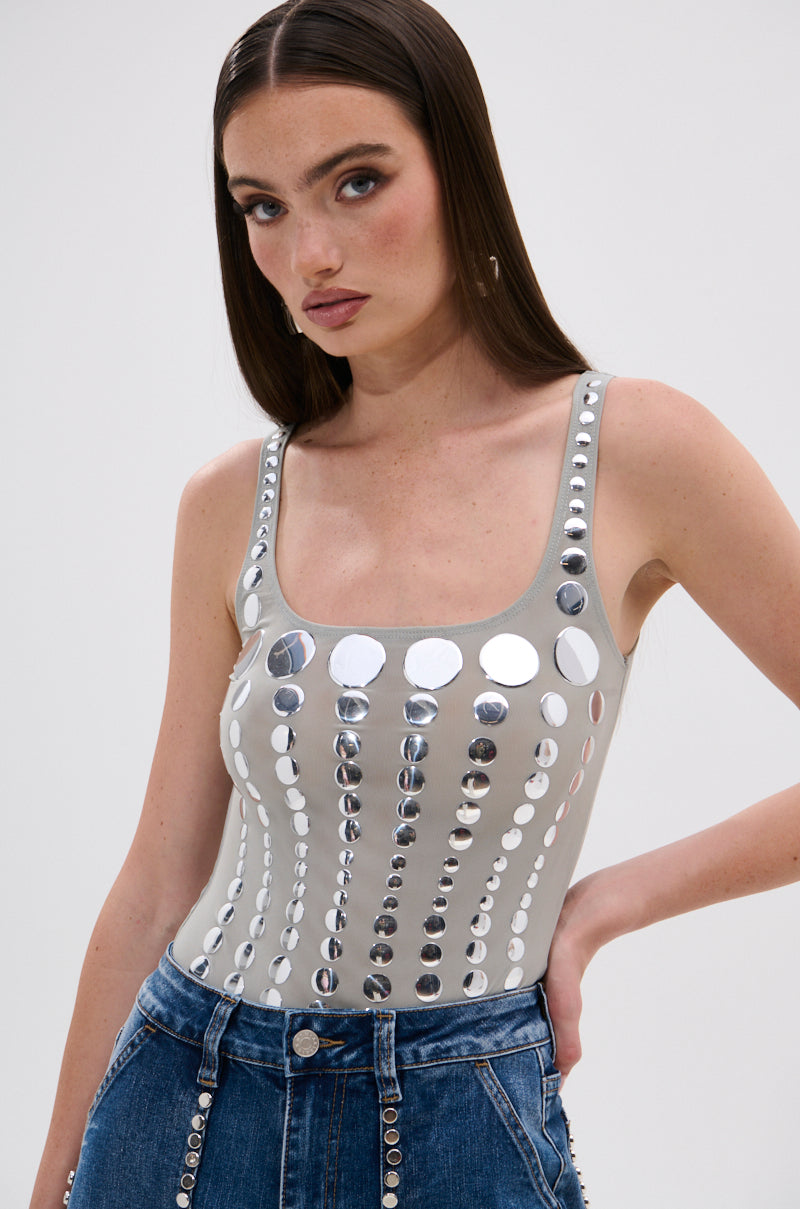 A woman with long brown hair wears the sleeveless, fitted ILLUSION POLKA DOT BODYSUIT with reflective mirrored disk embellishments and high-waisted blue jeans with button details, posing against a plain light background.