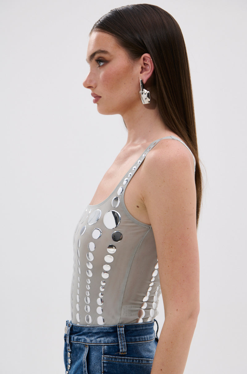 A woman with straight brown hair is shown in profile wearing the ILLUSION POLKA DOT BODYSUIT, featuring circular cut-outs and mirrored disk details, large abstract silver earrings, and blue jeans against a plain light background.
