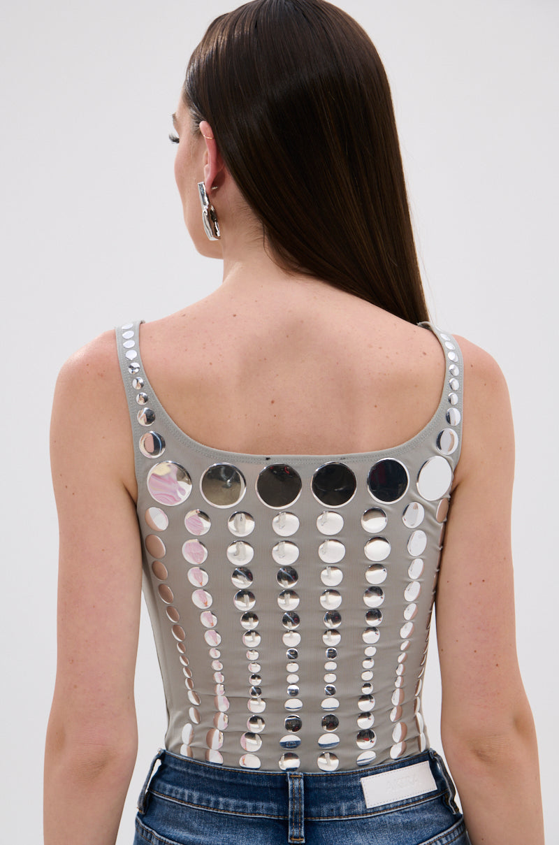 A woman with long brown hair is seen from behind, wearing the ILLUSION POLKA DOT BODYSUIT—a sleeveless, light gray stretchy mesh top with shiny silver dot details—and blue jeans.