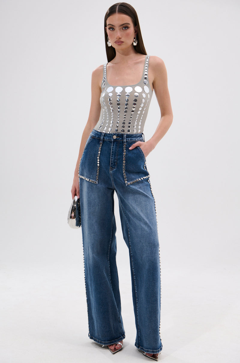A woman poses in the ILLUSION POLKA DOT BODYSUIT styled with high-waisted, wide-leg blue jeans. She holds a small handbag, accessorized with statement earrings and open-toe heels.