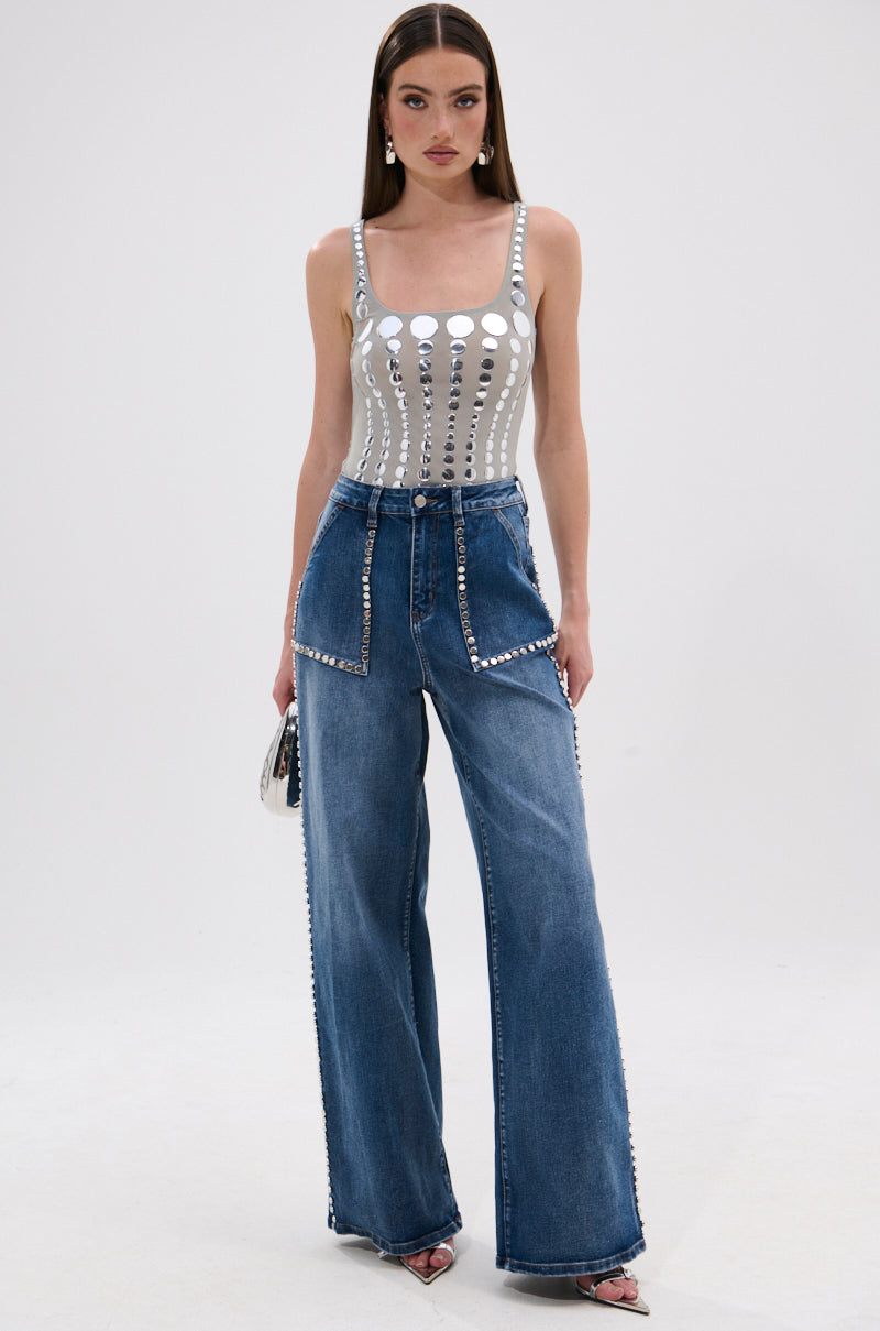 A woman poses against a white background wearing the ILLUSION POLKA DOT BODYSUIT, a silver metallic sleeveless top with mirrored disk embellishments, paired with high-waisted wide-leg blue jeans. She accessorizes with a small purse and heels.