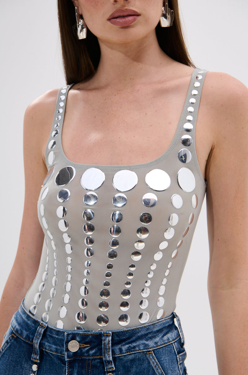 A woman wearing the ILLUSION POLKA DOT BODYSUIT with reflective mirrored disk embellishments, paired with blue denim jeans and large silver earrings.