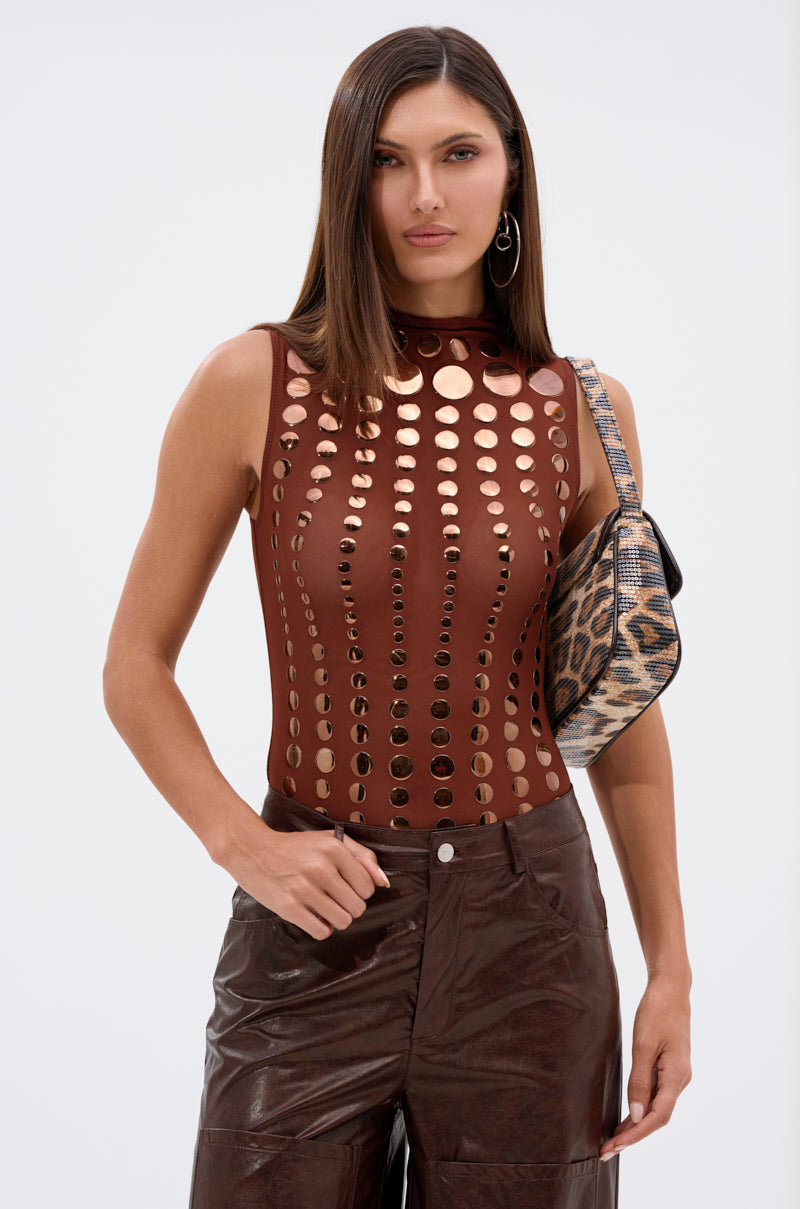 A woman stands against a light background wearing the NOT MY FAULT SLEEVELESS BODYSUIT IN BROWN, paired with brown leather pants and carrying a small leopard print shoulder bag.