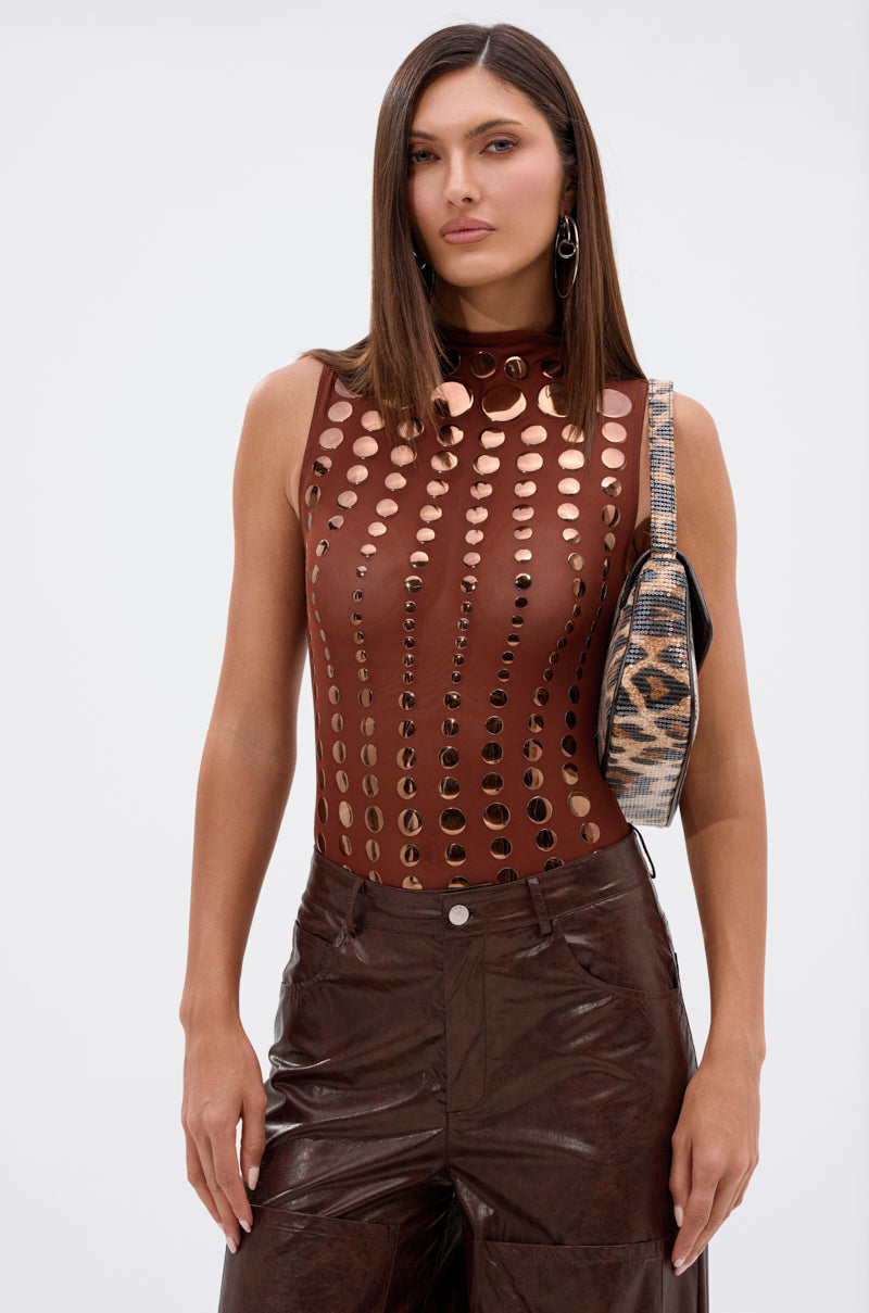 A woman with straight brown hair wears the NOT MY FAULT SLEEVELESS BODYSUIT IN BROWN, paired with dark brown leather pants and a patterned shoulder bag, standing against a plain light background.