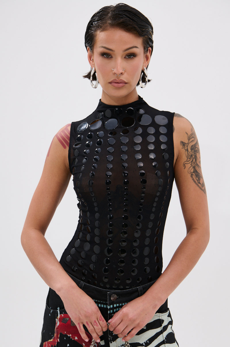 A woman with short dark hair wears the NOT MY FAULT SLEEVELESS BODYSUIT IN BLACK and patterned pants, accessorized with hoop earrings and arm tattoos, standing against a plain white background.