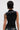 A person with short, dark, wet-look hair models the NOT MY FAULT SLEEVELESS BODYSUIT IN BLACK from the back, featuring cheeky circular cutouts, shiny accents, and revealing a detailed tattoo on their left arm.