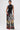 A woman poses against a white background wearing the NOT MY FAULT SLEEVELESS BODYSUIT IN BLACK, a semi-sheer mesh top with shiny dots, paired with high-waisted, wide-leg patchwork pants and a small silver purse.