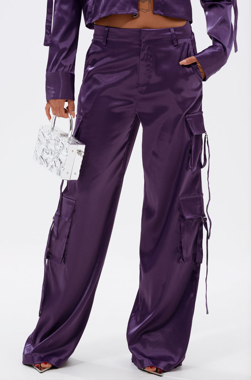 Someone wearing the MAYBE ITS YOU SATIN TROUSER in shiny purple satin with cargo-style pockets and a matching top, holding a small white handbag against a plain background. 1