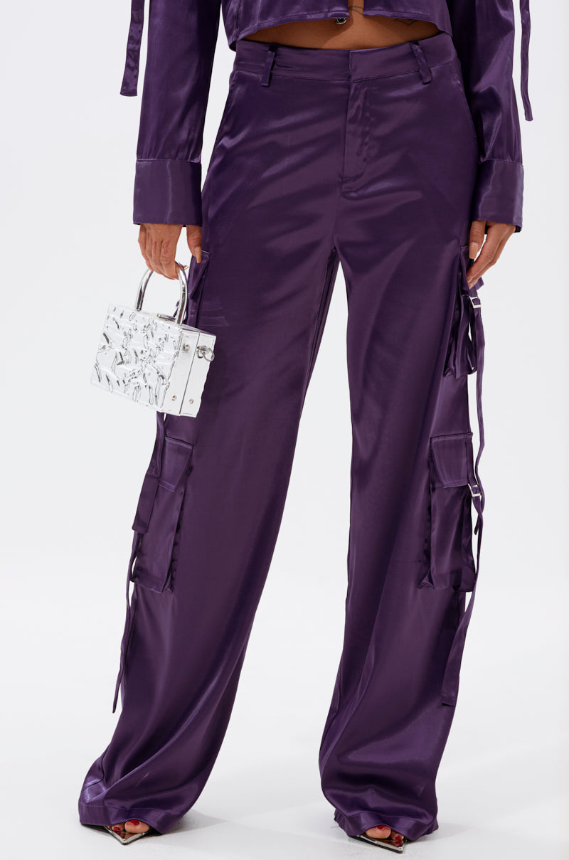 Wearing the MAYBE ITS YOU SATIN TROUSER in shiny purple with a high-rise fit and cargo pockets, a person holds a small silver metallic handbag while standing against a plain white background. 2