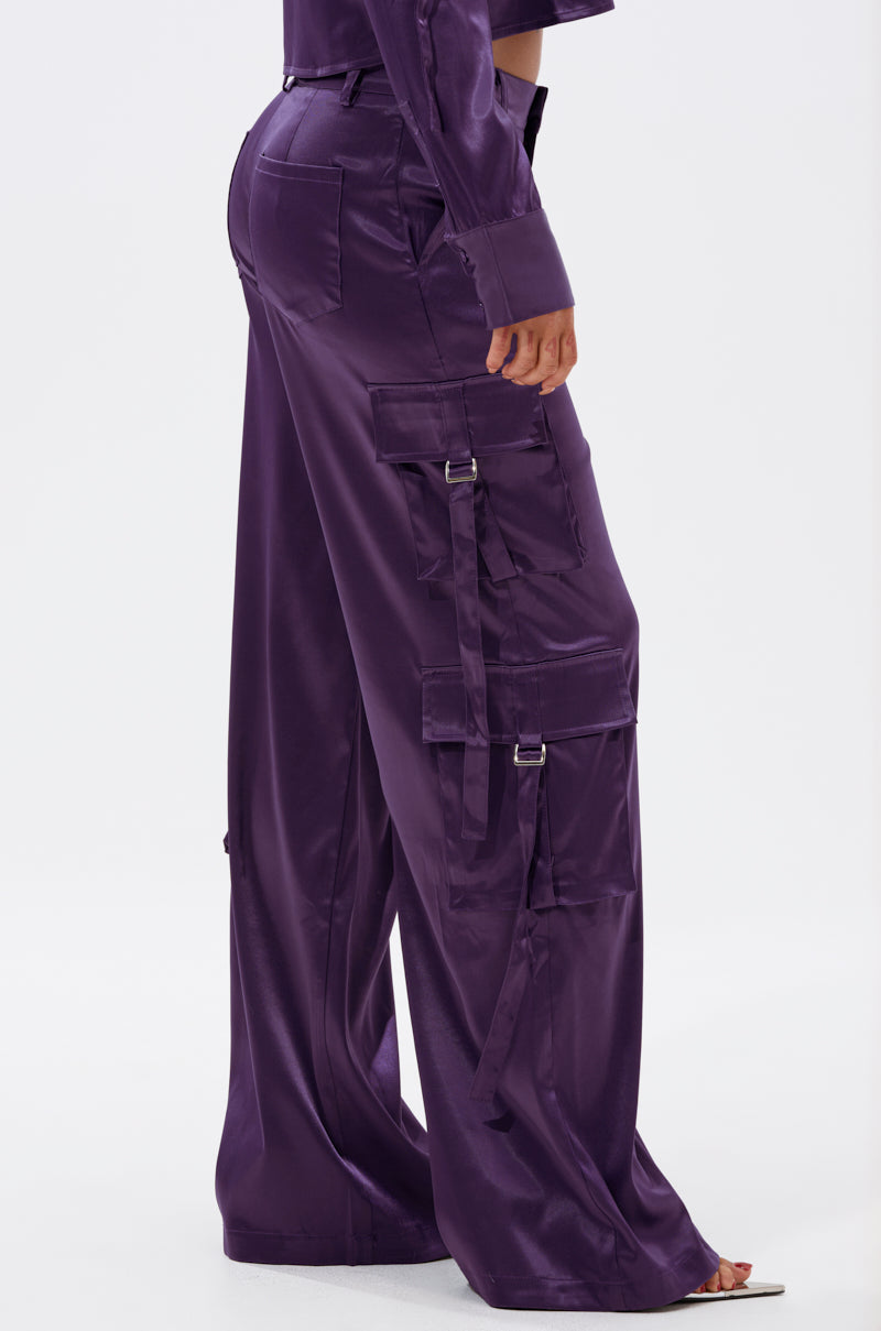 A person wearing the MAYBE ITS YOU SATIN TROUSER—high-rise, shiny purple satin with cargo-style pockets and buckle details—is shown from the waist down against a plain white background. 3