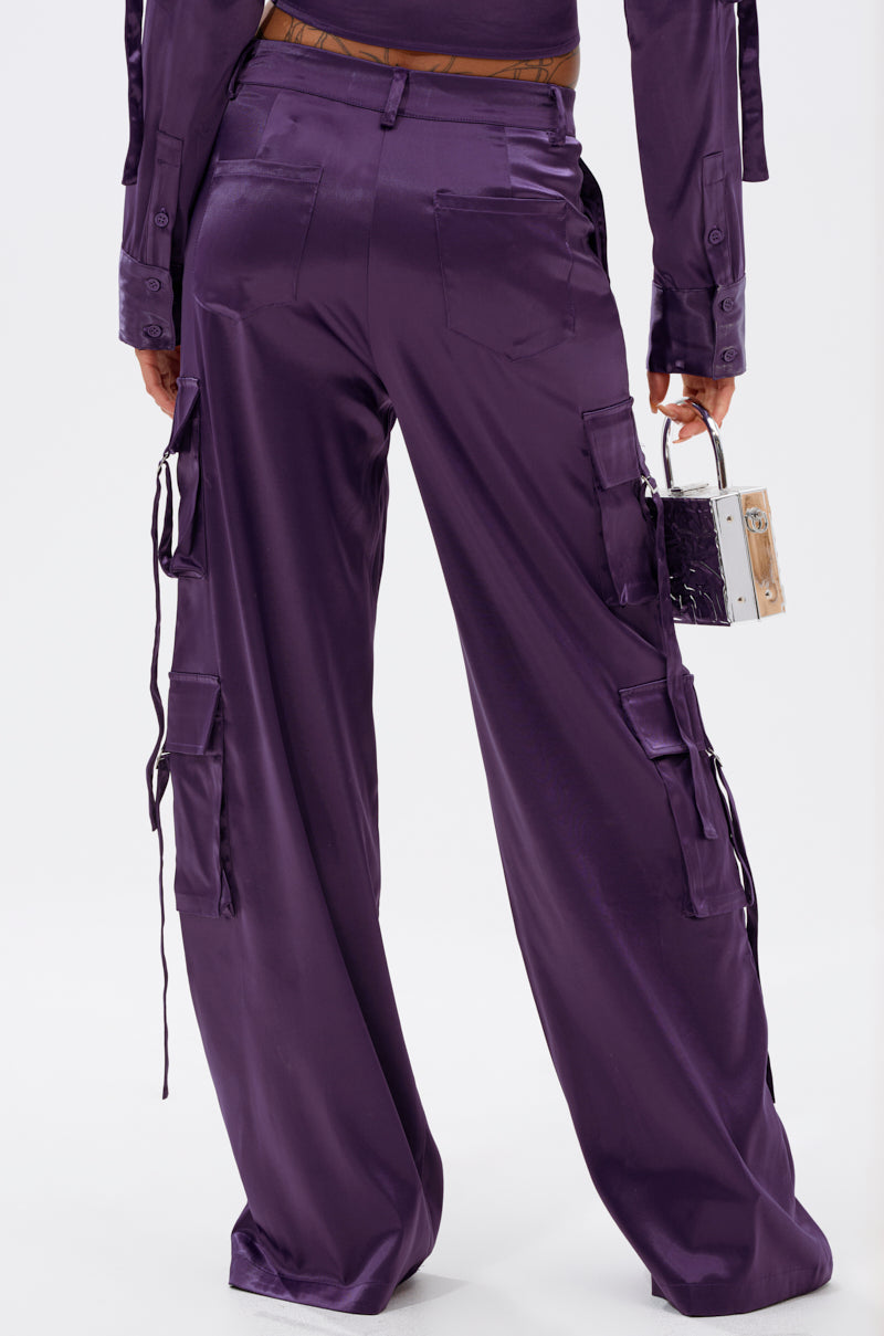 A person stands facing away, wearing loose-fitting MAYBE ITS YOU SATIN TROUSER in shiny purple with cargo pockets and ties, holding a small box-shaped handbag in their left hand. 4