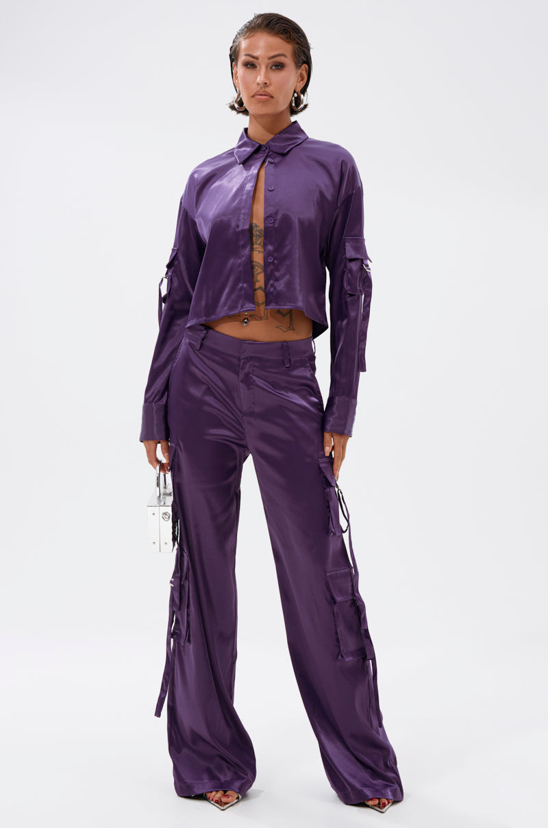 A woman poses against a white background wearing MAYBE ITS YOU SATIN TROUSER—high-rise, wide-leg, shiny purple satin pants with cargo pockets—paired with a matching cropped button-up shirt. She carries a small clear handbag and her hair is styled wet-look. 5