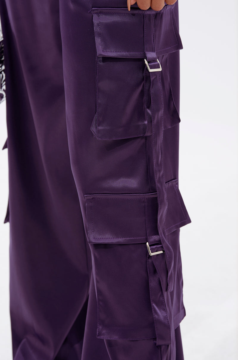A close-up shows a person wearing the MAYBE ITS YOU SATIN TROUSER in shiny purple with a high-rise fit and multiple cargo-style pockets featuring silver buckle details along the sides. The background is plain and light-colored. 6