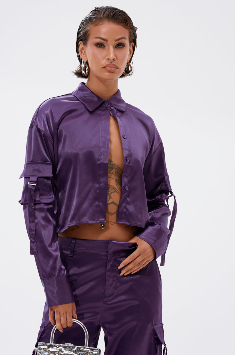 A woman with slicked-back hair wears the MAYBE ITS YOU SATIN BUTTON DOWN from AKIRA Label in shiny purple, styled partly unbuttoned with matching pants. She carries a small patterned handbag and shows a tattoo on her torso.