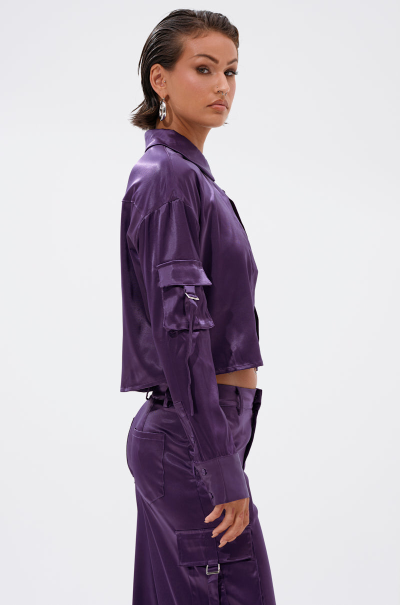 A woman with slicked-back hair models the MAYBE ITS YOU SATIN BUTTON DOWN, a shiny purple long-sleeve top with ruffle and zipper accents, paired with matching cargo-pocket pants. She stands sideways, facing the camera against a plain backdrop.