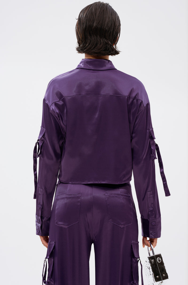 Person with short, dark hair wearing the MAYBE ITS YOU SATIN BUTTON DOWN in deep purple with matching cargo pants, seen from the back, holding a small clear box-shaped bag in their left hand.